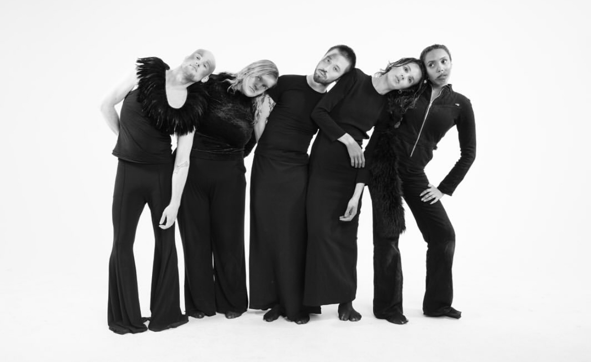 Five performers leaning into one another in black and white