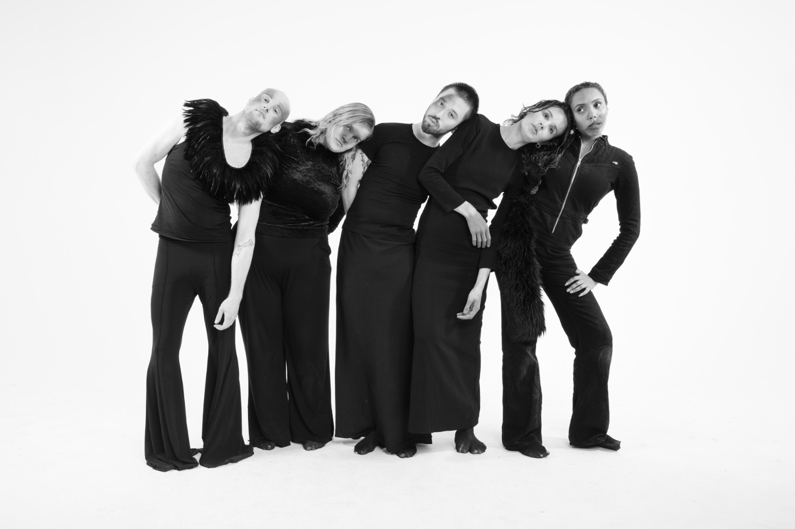 Five performers in black leaning together against a white studio background