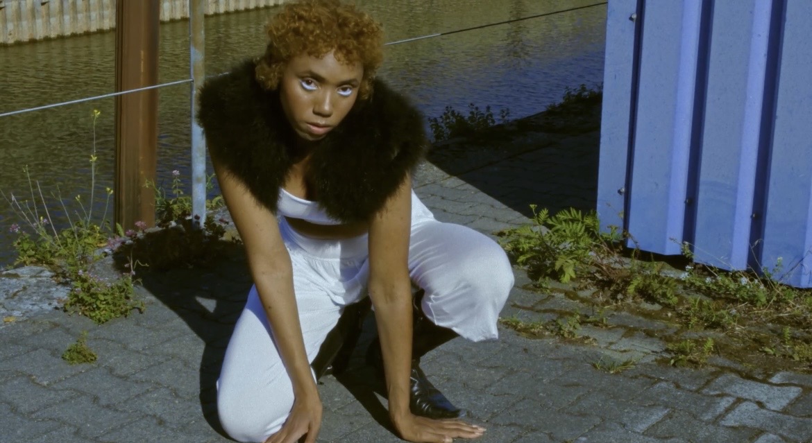Performer crouching beside a blue industrial structure near the water